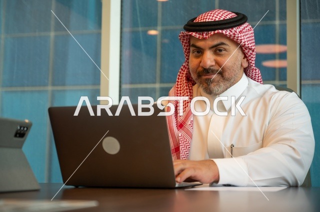 إيماءات الاندماج والتركيز، مهن وأعمال مكتبية، توفير بيئة عمل مريحة ومناسبة، استخدام جهاز الحاسوب المحمول لإنجاز المهام باحترافية، رجل عربي خليجي سعودي يرتدي الثوب والشماغ التقليدي يجلس في المكتب يقوم بالعمل عبر اللابتوب، التوظيف في المكاتب والشركات السعودية