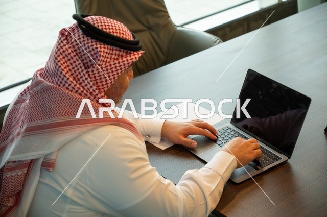 Employment in Saudi offices and companies, gestures of integration and focus, providing a comfortable and suitable work environment, using a laptop to accomplish tasks professionally, a background image of a Saudi Gulf Arab man wearing traditional thobe and ghutrah sitting at the office working on the laptop.