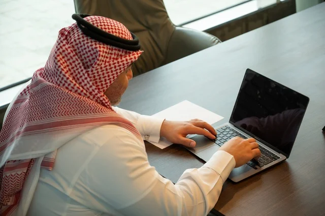 التوظيف في المكاتب والشركات السعودية، إيماءات الاندماج والتركيز، توفير بيئة عمل مريحة ومناسبة، استخدام جهاز الحاسوب المحمول لإنجاز المهام باحترافية، صورة خلفية لرجل عربي خليجي سعودي يرتدي الثوب والشماغ التقليدي يجلس في المكتب يقوم بالعمل عبر اللابتوب،