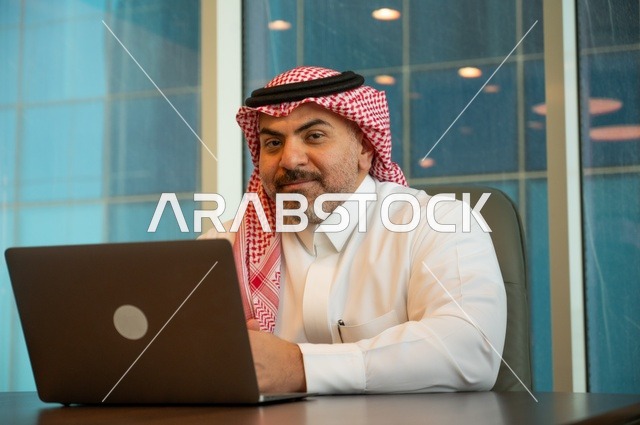 إيماءات الاندماج والتركيز، مهن وأعمال مكتبية، توفير بيئة عمل مريحة ومناسبة، استخدام جهاز الحاسوب المحمول لإنجاز المهام باحترافية، رجل عربي خليجي سعودي يرتدي الثوب والشماغ التقليدي يجلس في المكتب يقوم بالعمل عبر اللابتوب، التوظيف في المكاتب والشركات السعودية