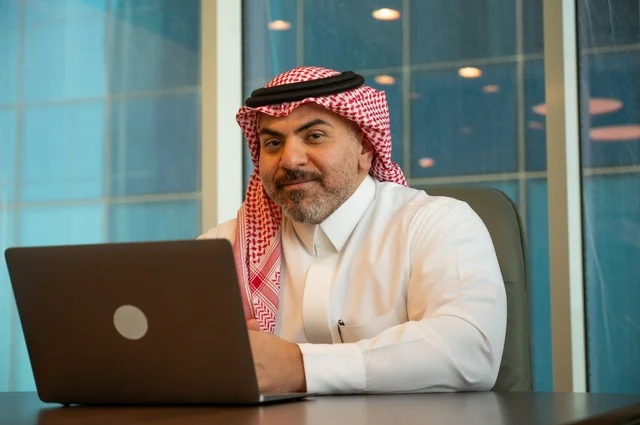 Gestures of integration and focus, professions and office work, providing a comfortable and suitable work environment, using a laptop to accomplish tasks professionally, an Arab Gulf Saudi man wearing traditional thobe and ghutrah sitting at the office working on the laptop, employment in Saudi offices and companies.
