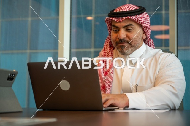 توفير بيئة عمل مريحة ومناسبة، التوظيف في المكاتب والشركات السعودية، إيماءات الاندماج والتركيز، استخدام جهاز الحاسوب المحمول لإنجاز المهام باحترافية، صورة خلفية لرجل عربي خليجي سعودي يرتدي الثوب والشماغ التقليدي يجلس في المكتب يقوم بالعمل عبر اللابتوب