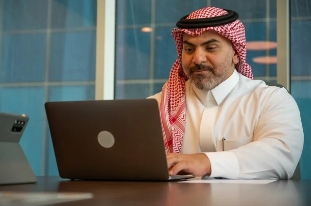 توفير بيئة عمل مريحة ومناسبة، التوظيف في المكاتب والشركات السعودية، إيماءات الاندماج والتركيز، استخدام جهاز الحاسوب المحمول لإنجاز المهام باحترافية، صورة خلفية لرجل عربي خليجي سعودي يرتدي الثوب والشماغ التقليدي يجلس في المكتب يقوم بالعمل عبر اللابتوب