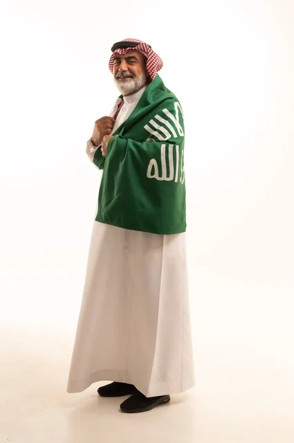 إيماءات الفخر و السرور بالانتماء للوطن, إحياء ذكرى اليوم الوطني السعودي 23 سبتمبر, بورتريه لرجل عربي خليجي سعودي مبتسم يرتدي الشماغ و الثوب التقليدي يحمل بيده علم يوم التأسيس، يوم بدينا 1727 م، الجسم بالطول الكامل، خلفية بيضاء