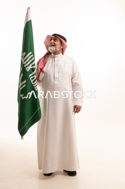 إحياء ذكرى اليوم الوطني السعودي 23 سبتمبر, إيماءات الفخر و السرور بالانتماء للوطن, بورتريه لرجل عربي خليجي سعودي مبتسم يرتدي الشماغ و الثوب التقليدي يحمل بيده علم يوم التأسيس، يوم بدينا 1727 م، الجسم بالطول الكامل، خلفية بيضاء