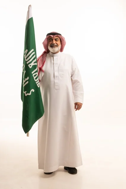 إحياء ذكرى اليوم الوطني السعودي 23 سبتمبر, إيماءات الفخر و السرور بالانتماء للوطن, بورتريه لرجل عربي خليجي سعودي مبتسم يرتدي الشماغ و الثوب التقليدي يحمل بيده علم يوم التأسيس، يوم بدينا 1727 م، الجسم بالطول الكامل، خلفية بيضاء