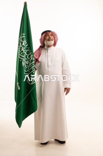 إحياء ذكرى اليوم الوطني السعودي 23 سبتمبر, إيماءات الفخر و السرور بالانتماء للوطن, بورتريه لرجل عربي خليجي سعودي مبتسم يرتدي الشماغ و الثوب التقليدي يحمل بيده علم يوم التأسيس، يوم بدينا 1727 م، الجسم بالطول الكامل، خلفية بيضاء