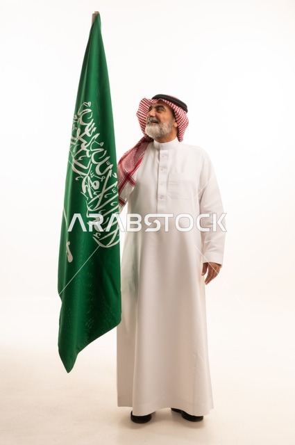 إحياء ذكرى اليوم الوطني السعودي 23 سبتمبر, إيماءات الفخر و السرور بالانتماء للوطن, بورتريه لرجل عربي خليجي سعودي مبتسم يرتدي الشماغ و الثوب التقليدي يحمل بيده علم يوم التأسيس، يوم بدينا 1727 م، الجسم بالطول الكامل، خلفية بيضاء