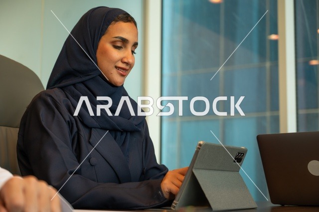 Integrating technology into work, remote business management, using a modern and advanced technical device, an Arab Gulf Saudi woman wearing an abaya and hijab using a tablet in a meeting room, completing required tasks via a laptop, a female office profession and job.