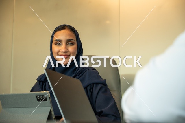 ايماءات السرور والبهجة، مهنة ووظيفة نسائية مكتبية ، دمج التكنولوجيا بالعمل، ادارة الأعمال عن بعد، استخدام جهاز تقني حديث ومتطور، امرأة عربية خليجية سعودية ترتدي العباءة والحجاب تقوم باستخدام الجهاز اللوحي في قاعة الاجتماع، إنجاز المهام المطلوبة عبر اللابتوب