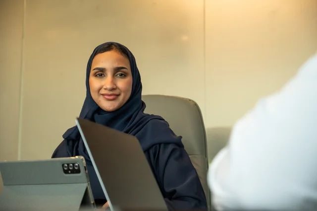 Gestures of joy and happiness, a profession and office job for women, integrating technology into work, remote business management, using a modern and advanced technical device, an Arab Gulf Saudi woman wearing an abaya and hijab using a tablet in the meeting room, completing the required tasks via laptop.
