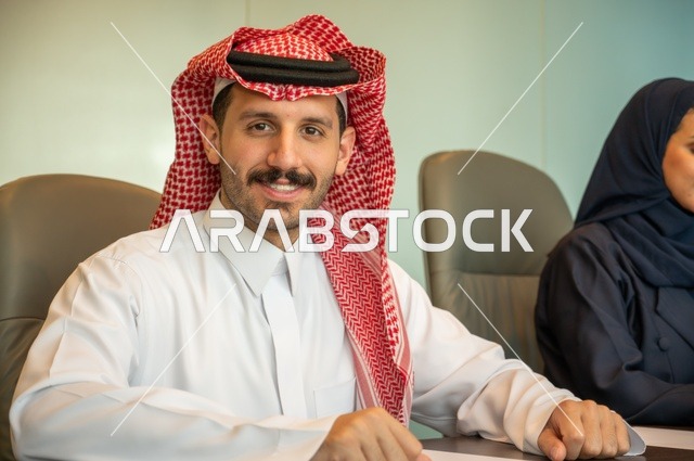إدارة شؤون الأعمال وتنظيمها، مهنة ووظيفة ادارية مكتبية، رجل عربي خليجي سعودي ينظر إلى الكاميرا بإيماءات سروروسعادة يرتدي الثوب التقليدي داخل قاعة الاجتماع، الشركات والمكاتب في المملكة العربية السعودية