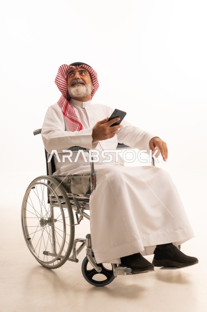التواصل مع الاهل والاصدقاء، تأمين اجهزة ومعدات تساعد بالحركة والتنقل، العنايه بأصحاب الهمم وتأمين احتياجاتهم الخاصة، بورتريه لرجل عربي خليجي سعودي من ذوي الاحتياجات الخاصة يرتدي الثوب التقليدي والشماغ يجلس على كرسي متحرك ويحمل بيده الهاتف المحمول، خلفية بيضاء