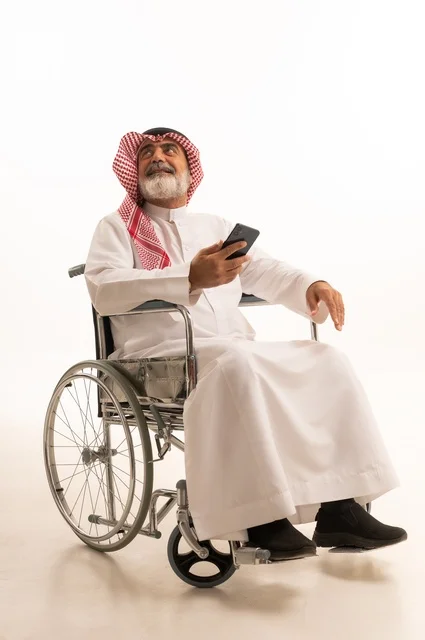 Communicating with family and friends, providing devices and equipment that help with movement and mobility, caring for people with disabilities and providing for their special needs, a portrait of a Saudi Arabian Gulf man with special needs wearing traditional thobe and shemagh sitting in a wheelchair and holding a mobile phone, white background