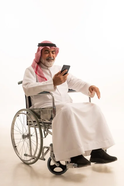 Communicating with family and friends, providing devices and equipment that help with movement and mobility, caring for people with disabilities and providing for their special needs, a portrait of a Saudi Arabian Gulf man with special needs wearing traditional thobe and shemagh sitting in a wheelchair and holding a mobile phone, white background