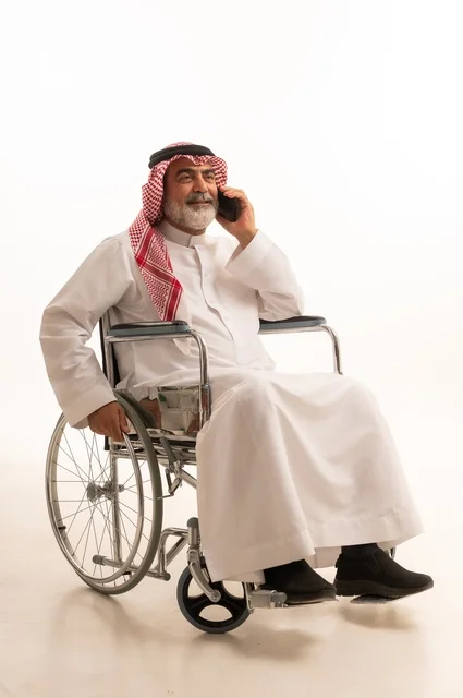 Making a phone call, providing mobility aids and equipment, caring for people with disabilities and providing for their special needs, portrait of a Saudi Arabian Gulf man with special needs wearing traditional thobe and shemagh sitting in a wheelchair and holding a mobile phone, white background