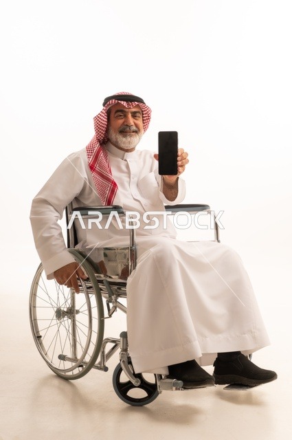 عرض شاشة سوداء فارغة، تأمين اجهزة ومعدات تساعد بالحركة والتنقل، العنايه بأصحاب الهمم وتأمين احتياجاتهم الخاصة، بورتريه لرجل عربي خليجي سعودي من ذوي الاحتياجات الخاصة يرتدي الثوب التقليدي والشماغ يجلس على كرسي متحرك ويحمل بيده الهاتف المحمول، خلفية بيضاء