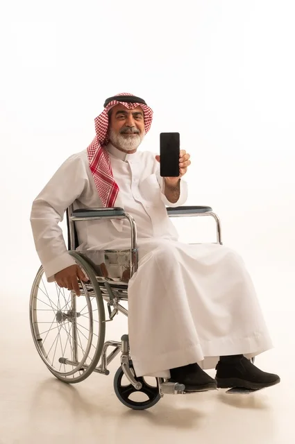 عرض شاشة سوداء فارغة، تأمين اجهزة ومعدات تساعد بالحركة والتنقل، العنايه بأصحاب الهمم وتأمين احتياجاتهم الخاصة، بورتريه لرجل عربي خليجي سعودي من ذوي الاحتياجات الخاصة يرتدي الثوب التقليدي والشماغ يجلس على كرسي متحرك ويحمل بيده الهاتف المحمول، خلفية بيضاء