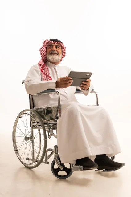 الاستفادة من التقنيات الحديثة والتكنولوجيا المتطورة، تأمين اجهزة ومعدات تساعد بالحركة والتنقل، ضعف القدرة الحركية، بورتريه لرجل عربي خليجي سعودي من ذوي الاحتياجات الخاصة يرتدي الثوب التقليدي والشماغ يجلس على كرسي متحرك ويحمل بيده الجهاز اللوحي، خلفية بيضاء