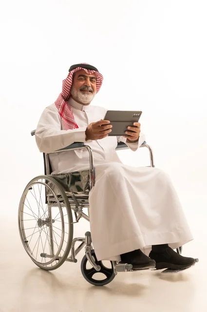 الاستفادة من التقنيات الحديثة والتكنولوجيا المتطورة، تأمين اجهزة ومعدات تساعد بالحركة والتنقل، ضعف القدرة الحركية، بورتريه لرجل عربي خليجي سعودي من ذوي الاحتياجات الخاصة يرتدي الثوب التقليدي والشماغ يجلس على كرسي متحرك ويحمل بيده الجهاز اللوحي، خلفية بيضاء