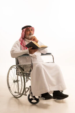 Spending free time in education and learning, developing reading skills, interest in browsing useful books, poor motor ability and difficulty walking, portrait of a Saudi Arabian Gulf man with special needs wearing traditional thobe and shemagh sitting in a wheelchair and holding a book in his hand, white background