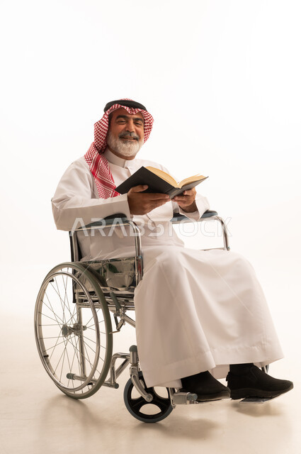 Spending free time in education and learning, developing reading skills, interest in browsing useful books, poor motor ability and difficulty walking, portrait of a Saudi Arabian Gulf man with special needs wearing traditional thobe and shemagh sitting in a wheelchair and holding a book in his hand, white background