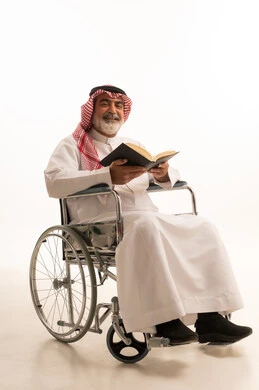 Spending free time in education and learning, developing reading skills, interest in browsing useful books, poor motor ability and difficulty walking, portrait of a Saudi Arabian Gulf man with special needs wearing traditional thobe and shemagh sitting in a wheelchair and holding a book in his hand, white background