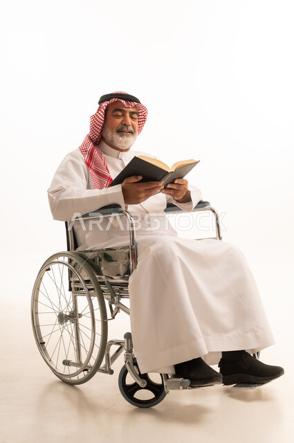 قضاء أوقات الفراغ بالتثقيف والتعلم, تنمية مهارات القراءة, الاهتمام بتصفح الكتب المفيدة، ضعف القدرة الحركية وصعوبة المشي، بورتريه لرجل عربي خليجي سعودي من ذوي الاحتياجات الخاصة يرتدي الثوب التقليدي والشماغ يجلس على كرسي متحرك ويحمل بيده كتاب، خلفية بيضاء