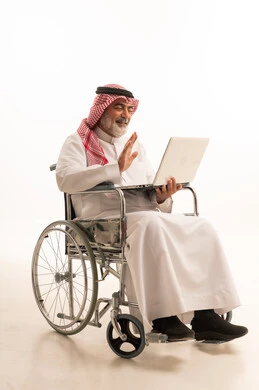 العمل عن بعد، العنايه بأصحاب الهمم وتأمين احتياجاتهم الخاصة، ضعف القدرة الحركية وصعوبة المشي، بورتريه لرجل عربي خليجي سعودي من ذوي الاحتياجات الخاصة يرتدي الثوب التقليدي والشماغ يجلس على كرسي متحرك ويحمل بيده الحاسوب المحمول، خلفية بيضاء