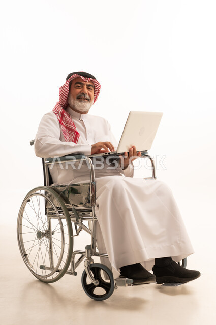 Remote work, caring for people with disabilities and providing for their special needs, poor motor ability and difficulty walking, portrait of a Saudi Arabian Gulf man with special needs wearing traditional thobe and shemagh sitting in a wheelchair and holding a laptop in his hand, white background