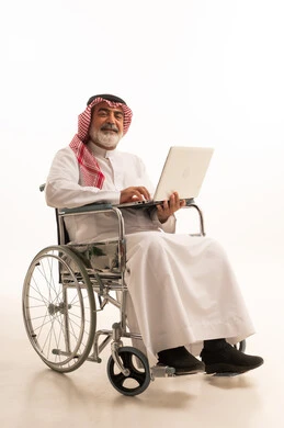 العمل عن بعد، العنايه بأصحاب الهمم وتأمين احتياجاتهم الخاصة، ضعف القدرة الحركية وصعوبة المشي، بورتريه لرجل عربي خليجي سعودي من ذوي الاحتياجات الخاصة يرتدي الثوب التقليدي والشماغ يجلس على كرسي متحرك ويحمل بيده الحاسوب المحمول، خلفية بيضاء