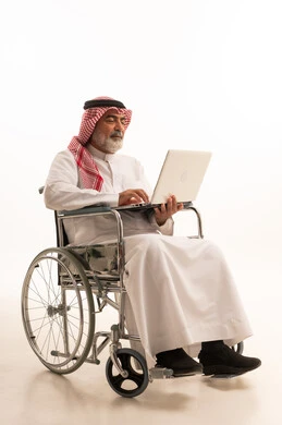 Remote work, caring for people with disabilities and providing for their special needs, poor motor ability and difficulty walking, portrait of a Saudi Arabian Gulf man with special needs wearing traditional thobe and shemagh sitting in a wheelchair and holding a laptop in his hand, white background