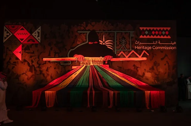 The Heritage Authority is a Saudi Arabian Arab organization established in 2020 as part of the Ministry of Culture. Its goal is to protect, preserve, and develop the cultural heritage of the Kingdom of Saudi Arabia. A poster in an exhibition features a Gulf Arab Saudi woman weaving colorful threads.
