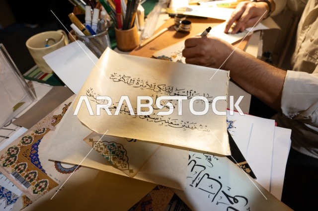 رجل يمارس فن الخط العربي، خطاط منهمك في كتابة لوحات خطية باستخدام أدوات الخط التقليدية ،أقلام، حبر، أعمال خطية جاهزة أو قيد التنفيذ