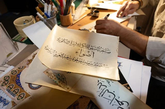 رجل يمارس فن الخط العربي، خطاط منهمك في كتابة لوحات خطية باستخدام أدوات الخط التقليدية ،أقلام، حبر، أعمال خطية جاهزة أو قيد التنفيذ