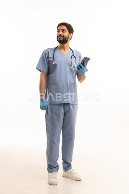 تقديم الخدمات الصحية للمرضى في المراكز والمستشفيات، مفهوم الطب والرعاية الصحية، بورتريه لممرض عربي خليجي سعودي يرتدي اليونيفورم مع السماعة الطبية يحمل بيده الهاتف المحمول، مهنة ووظيفة طبية، العمل في القطاع الصحي في المملكة، الجسم بالطول الكامل، خلفيه بيضاء