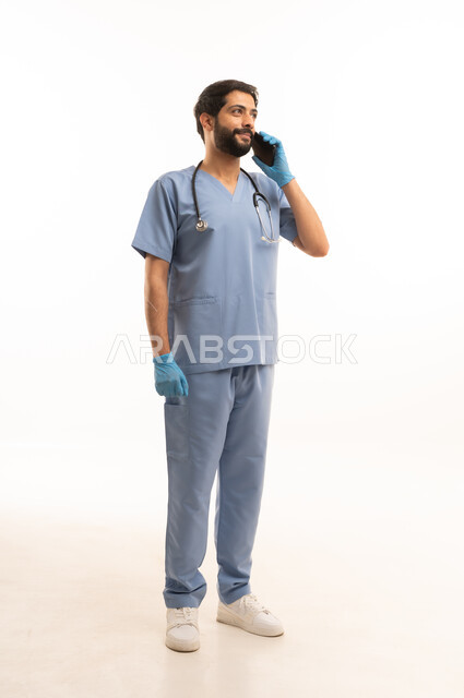 اجراء مكالمة هاتفية، تقديم الخدمات الصحية للمرضى، مفهوم الطب والرعاية الصحية، بورتريه لممرض عربي خليجي سعودي يرتدي اليونيفورم مع السماعة الطبية يحمل بيده الهاتف المحمول، مهنة ووظيفة طبية، العمل في القطاع الصحي في المملكة، الجسم بالطول الكامل، خلفيه بيضاء