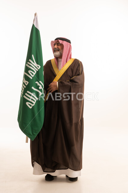 الاحتفال بذكرى اليوم الوطني السعودي 23 سبتمبر, إيماءات الفخر و السرور بالانتماء للوطن, بورتريه لرجل عربي خليجي سعودي مبتسم يرتدي الشماغ و الثوب التقليدي مع البشت يحمل بيده علم يوم التأسيس، يوم بدينا 1727 م، الجسم بالطول الكامل، خلفية بيضاء