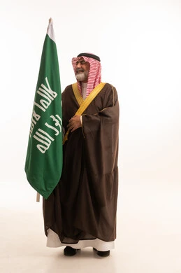Celebrating Saudi National Day, September 23, gestures of pride and joy in belonging to the homeland, portrait of a smiling Saudi Arabian Gulf man wearing a traditional shemagh and thobe with a bisht holding the Founding Day flag, the day we began 1727 AD, full body, white background