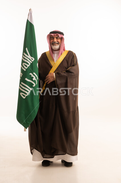 الاحتفال بذكرى اليوم الوطني السعودي 23 سبتمبر, إيماءات الفخر و السرور بالانتماء للوطن, بورتريه لرجل عربي خليجي سعودي مبتسم يرتدي الشماغ و الثوب التقليدي مع البشت يحمل بيده علم يوم التأسيس، يوم بدينا 1727 م، الجسم بالطول الكامل، خلفية بيضاء