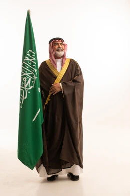 Celebrating Saudi National Day, September 23, gestures of pride and joy in belonging to the homeland, portrait of a smiling Saudi Arabian Gulf man wearing a traditional shemagh and thobe with a bisht holding the Founding Day flag, the day we began 1727 AD, full body, white background