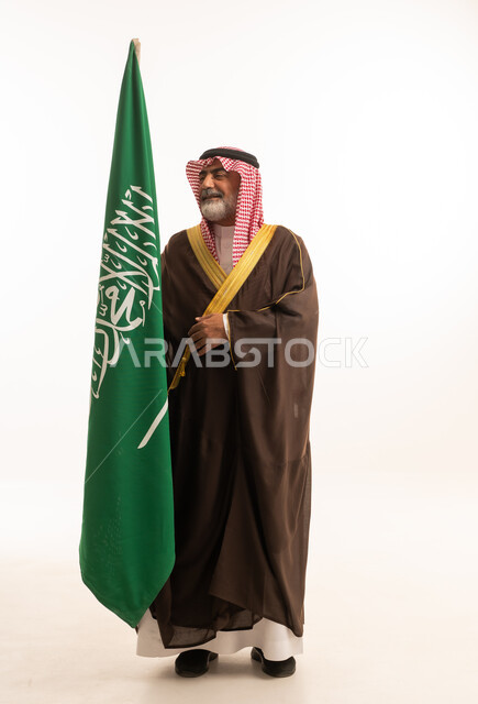 الاحتفال بذكرى اليوم الوطني السعودي 23 سبتمبر, إيماءات الفخر و السرور بالانتماء للوطن, بورتريه لرجل عربي خليجي سعودي مبتسم يرتدي الشماغ و الثوب التقليدي مع البشت يحمل بيده علم يوم التأسيس، يوم بدينا 1727 م، الجسم بالطول الكامل، خلفية بيضاء