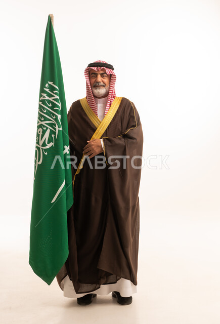 الاحتفال بذكرى اليوم الوطني السعودي 23 سبتمبر, إيماءات الفخر و السرور بالانتماء للوطن, بورتريه لرجل عربي خليجي سعودي مبتسم يرتدي الشماغ و الثوب التقليدي مع البشت يحمل بيده علم يوم التأسيس، يوم بدينا 1727 م، الجسم بالطول الكامل، خلفية بيضاء