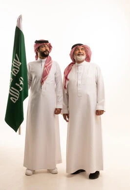  إيماءات الفخر و السرور بالانتماء للوطن, الاحتفال بذكرى اليوم الوطني السعودي 23 سبتمبر, بورتريه لرجل عربي خليجي سعودي مبتسم يرتدي الشماغ و الثوب التقليدي مع البشت يحمل بيده علم يوم التأسيس ويقف مع والده، يوم بدينا 1727 م، الجسم بالطول الكامل، خلفية بيضاء