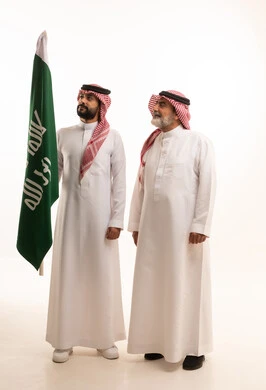  إيماءات الفخر و السرور بالانتماء للوطن, الاحتفال بذكرى اليوم الوطني السعودي 23 سبتمبر, بورتريه لرجل عربي خليجي سعودي مبتسم يرتدي الشماغ و الثوب التقليدي مع البشت يحمل بيده علم يوم التأسيس ويقف مع والده، يوم بدينا 1727 م، الجسم بالطول الكامل، خلفية بيضاء