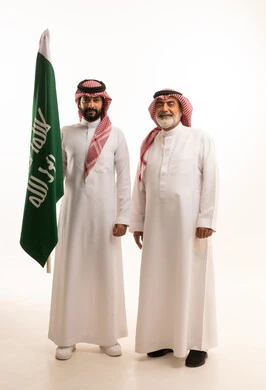 Gestures of pride and joy in belonging to the homeland, celebrating the Saudi National Day on September 23, portrait of a smiling Saudi Arabian Gulf man wearing a traditional shemagh and thobe with a bisht holding the Founding Day flag and standing with his father, the day we began 1727 AD, full body, white background