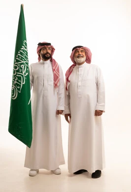 Gestures of pride and joy in belonging to the homeland, celebrating the Saudi National Day on September 23, portrait of a smiling Saudi Arabian Gulf man wearing a traditional shemagh and thobe with a bisht holding the Founding Day flag and standing with his father, the day we began 1727 AD, full body, white background