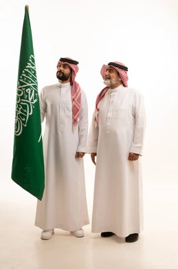  إيماءات الفخر و السرور بالانتماء للوطن, الاحتفال بذكرى اليوم الوطني السعودي 23 سبتمبر, بورتريه لرجل عربي خليجي سعودي مبتسم يرتدي الشماغ و الثوب التقليدي مع البشت يحمل بيده علم يوم التأسيس ويقف مع والده، يوم بدينا 1727 م، الجسم بالطول الكامل، خلفية بيضاء