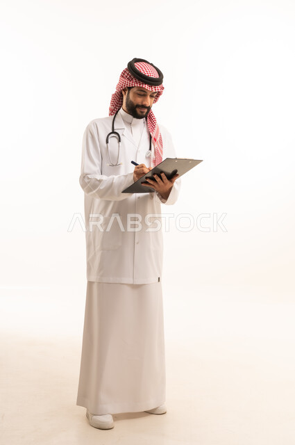 تدوين وتسجيل المعلومات، مفهوم الطب والرعاية الصحية، بورتريه لطبيب عربي خليجي سعودي يرتدي الثوب التقليدي مع المعطف والسماعة يقدم الخدمات الصحية للمرضى في المراكز والمستشفيات، مهنة ووظيفة طبية، العمل في القطاع الصحي في المملكة، الجسم بالطول الكامل، خلفيه بيضاء