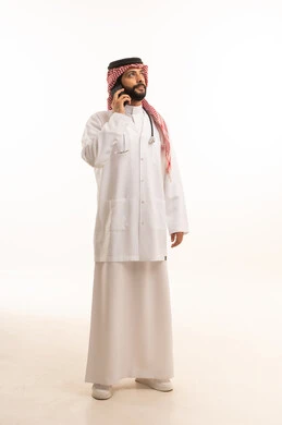Making a phone call on a mobile phone, concept of medicine and healthcare, portrait of a Saudi Arabian Gulf doctor wearing traditional thobe with coat and stethoscope providing health services to patients, medical profession and job, working in the health sector in the Kingdom, full body, white background
